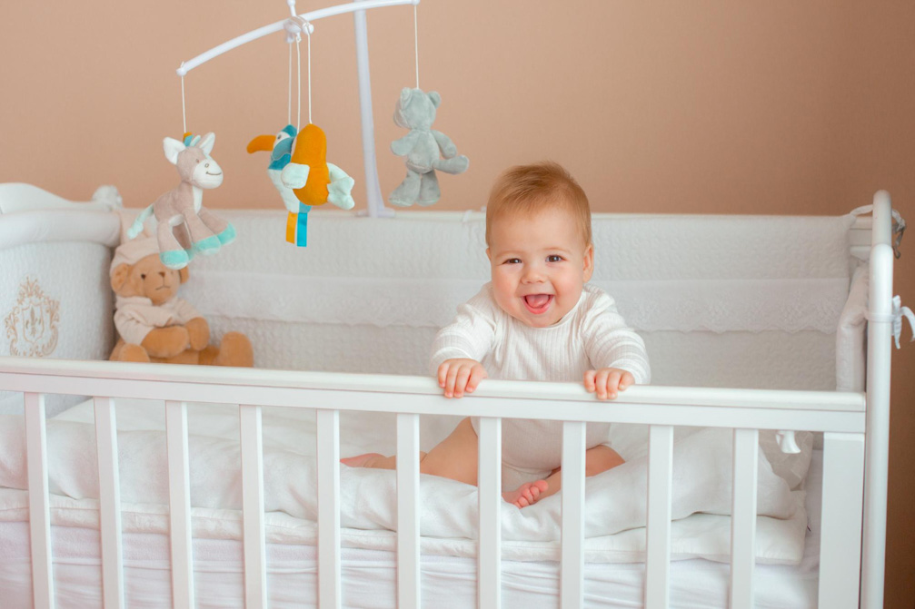 Χαμογελαστό μωρό κάθεται μέσα στη βρεφική του κούνια, πιάνοντας με τα χέρια του τα προστατευτικά του κρεβατιού.