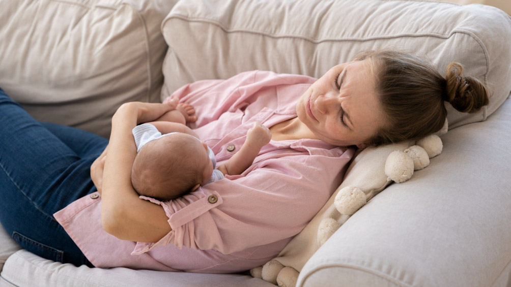 Νέα γυναίκα ξαπλωμένη στον καναπέ κρατάει αγκαλιά το νεογέννητο βρέφος της.