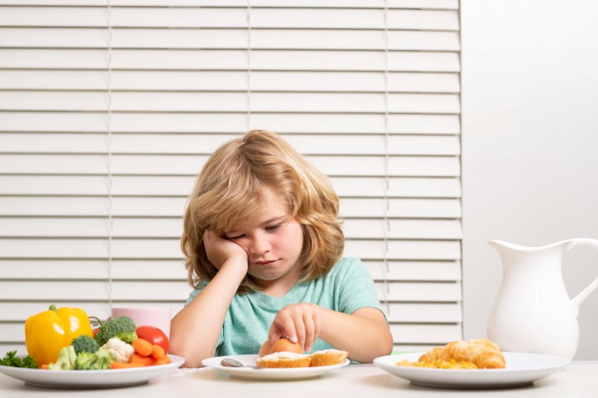 Μικρό παιδί κάθεται στο τραπέζι και κοιτάει χωρίς όρεξη τα υγιεινά φαγητά πάνω σε αυτό.
